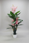 A potted artificial plant with glossy green leaves and pink-and-white flowers, standing in a white plastic pot on a light wood floor against a plain wall.