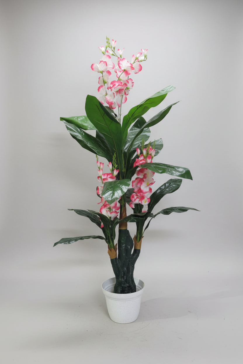 A potted artificial plant with glossy green leaves and pink-and-white flowers, standing in a white plastic pot on a light wood floor against a plain wall.